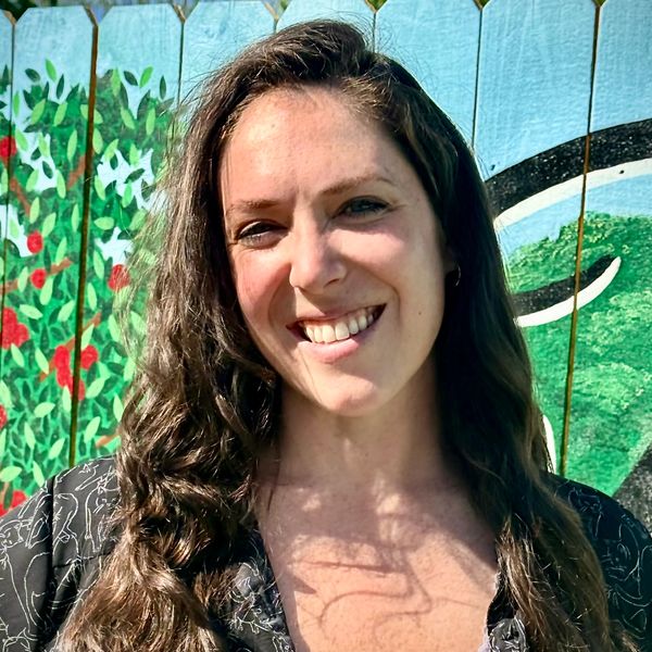 Smiling woman with long curly hair in front of a colorful painted fence.