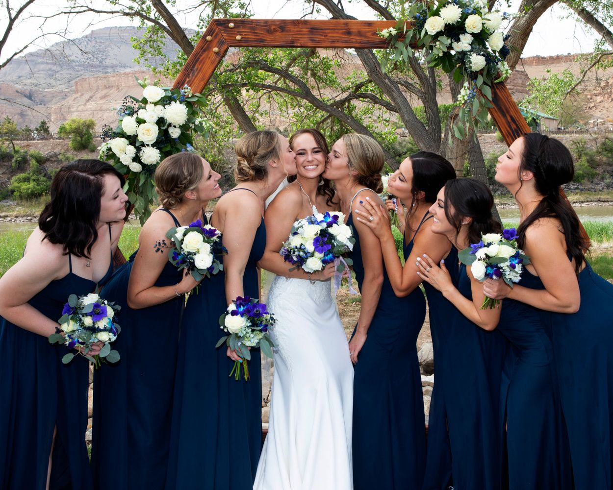 Bride in white dress kissed by bridesmaids in navy blue dresses outdoors.