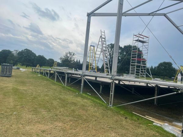 Outdoor stage construction with ladders and scaffolding on grassy field.