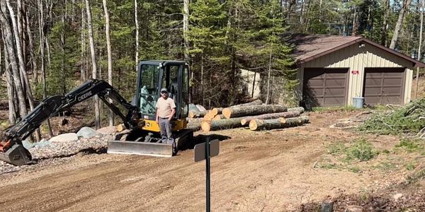 Man operating excavator near wood logs and garage in wooded area.