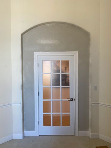White glass-paneled door set in an arched gray wall section inside a beige room.
