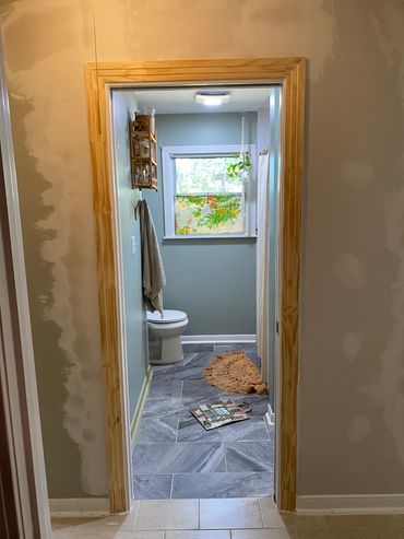 View into a bathroom with gray tiles, a toilet, and a window with a floral curtain.