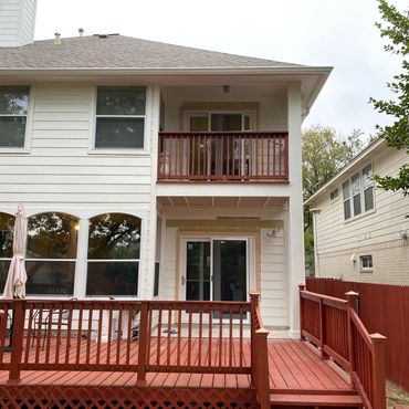 Two-story white house with red wooden deck and balcony under cloudy sky.