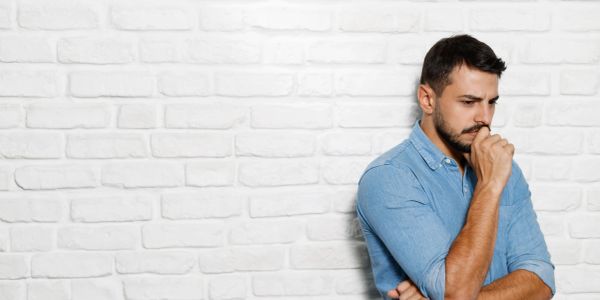 A man in a blue shirt looks deep in thought against a white brick wall.