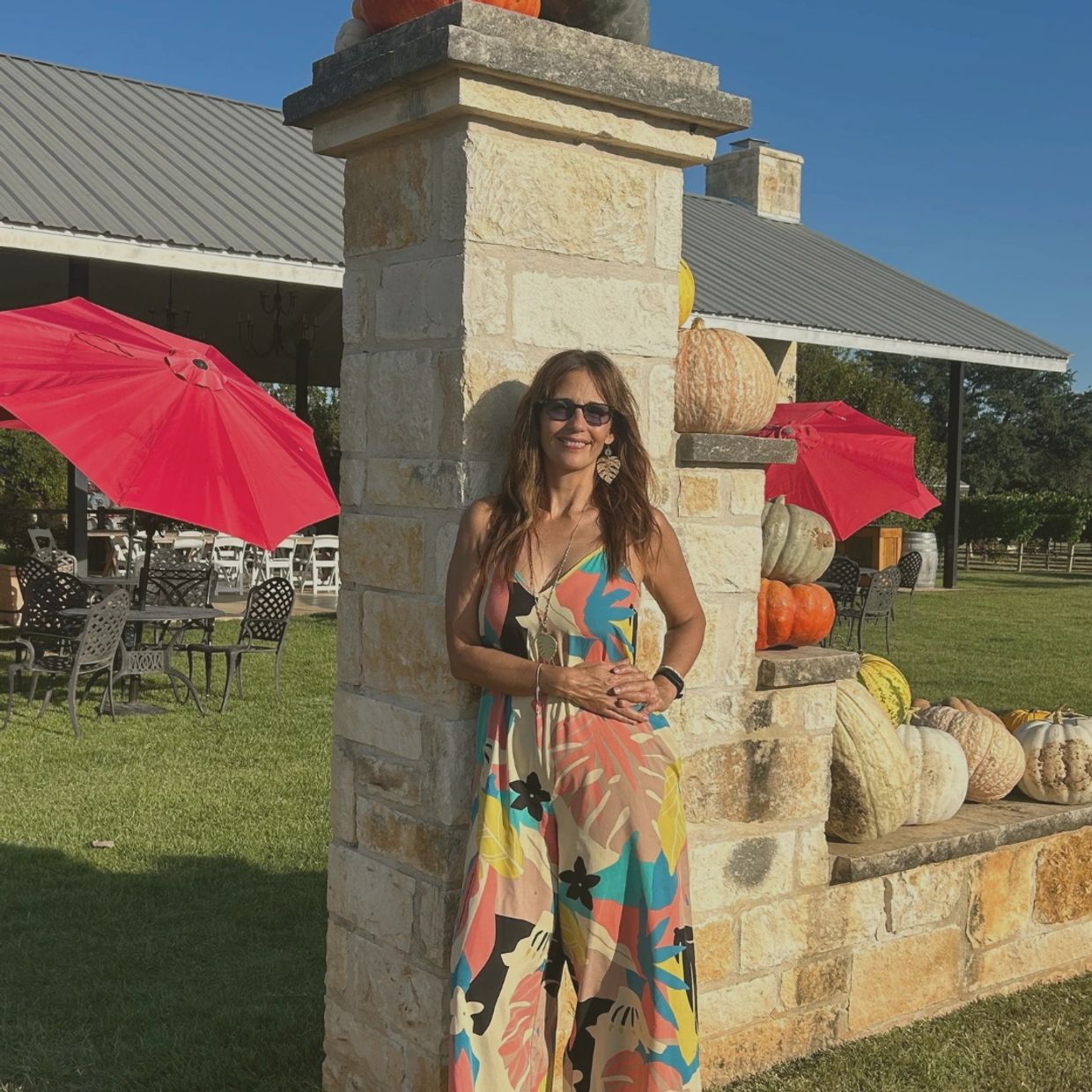 Woman in colorful dress stands by stone pillar decorated with pumpkins and red umbrellas.