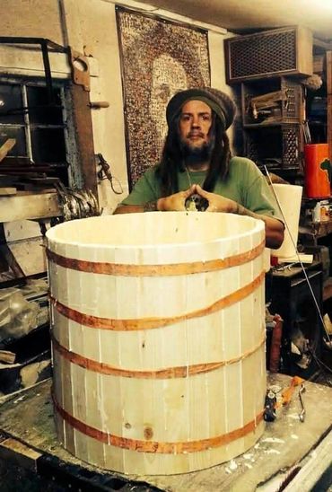 Man with dreadlocks sitting behind a large wooden barrel in a workshop.
