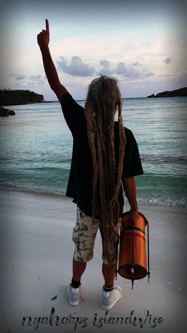 Man with long dreadlocks holding a drum, raising one arm on a beach at sunset.