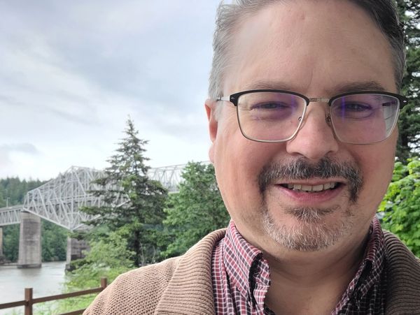 Smiling man with glasses outdoors near a river and bridge.