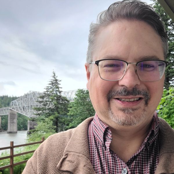 Smiling man with glasses outdoors near a river and bridge.