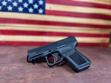 Black handgun on wooden surface with American flag backdrop.