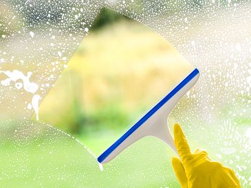 A yellow-gloved hand using a squeegee to clean a soapy window.