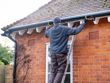 Worker on a ladder cleaning or inspecting house gutters on a brick home with a mossy roof.