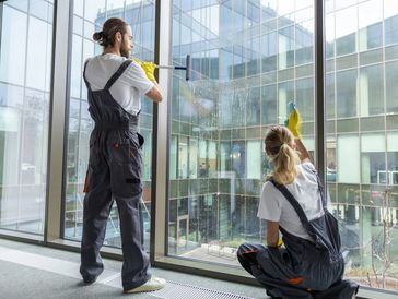 Two professional window cleaners in uniforms cleaning large glass windows in a modern building.