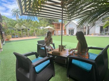 Two women relaxing with drinks under a striped umbrella in a tropical garden setting.