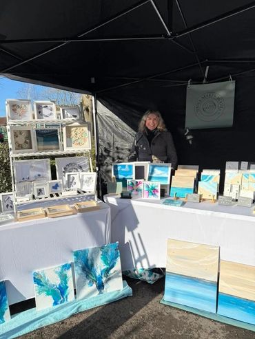 Artist showcasing vibrant blue and beige abstract paintings at an outdoor market stall.