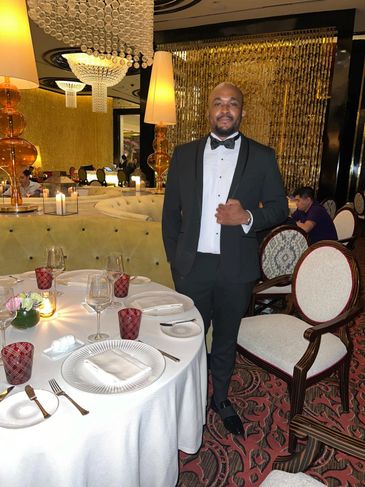 Man in a black tuxedo standing next to a formal dining table in an elegant restaurant.
