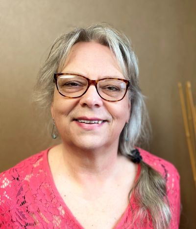 A woman with long, gray hair, wearing a pink top and eyeglasses