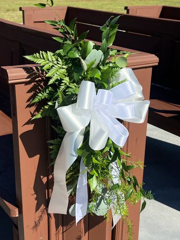 White ribbon and greenery decoration on a wooden pew outdoors.