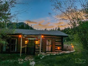 A rustic log cabin at dusk with lights glowing near the door and along the path on the side. The sky