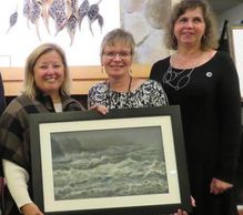 Three women smiling, holding a framed seascape painting together.