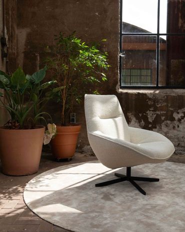 De Gigi fauteuil van Cartel Living in een lichte stof op een rond tapijt en op de achtergrond plante