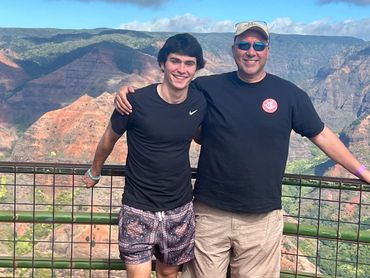 Two men smiling with scenic mountain backdrop under blue sky.