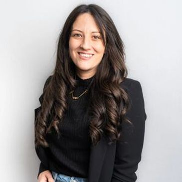 Smiling woman with long curly hair wearing a black blazer and jeans.