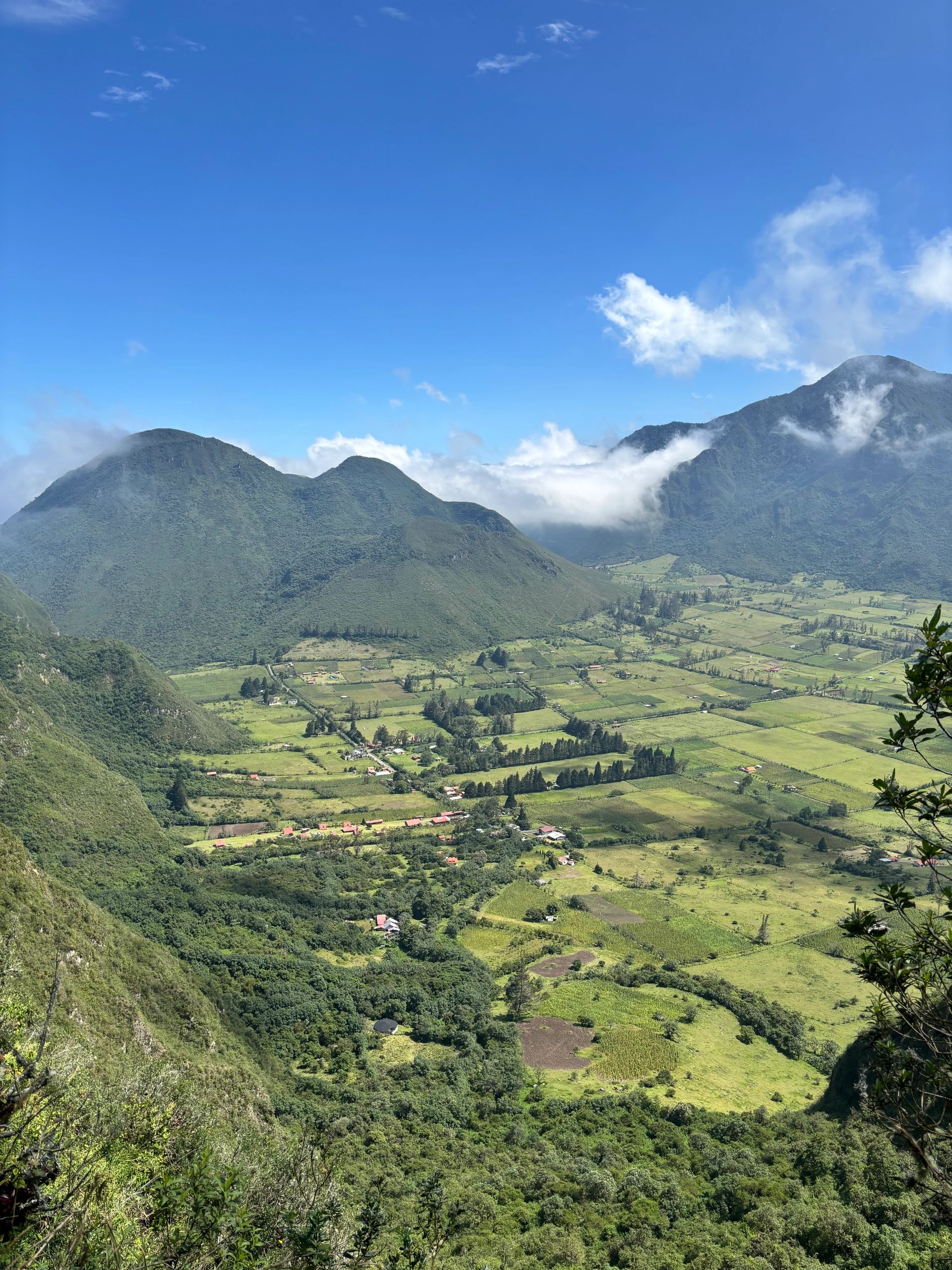 Lush green valley with mountains and scattered clouds under a clear blue sky.