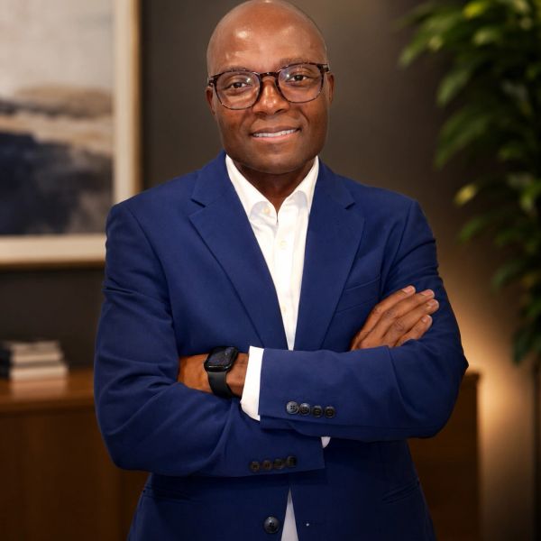 Confident man in a blue suit with glasses, arms crossed, smiling.