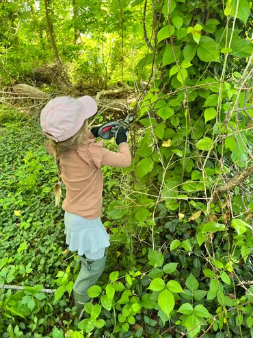 Child cutting vines in a dense green forest wearing gloves and boots.