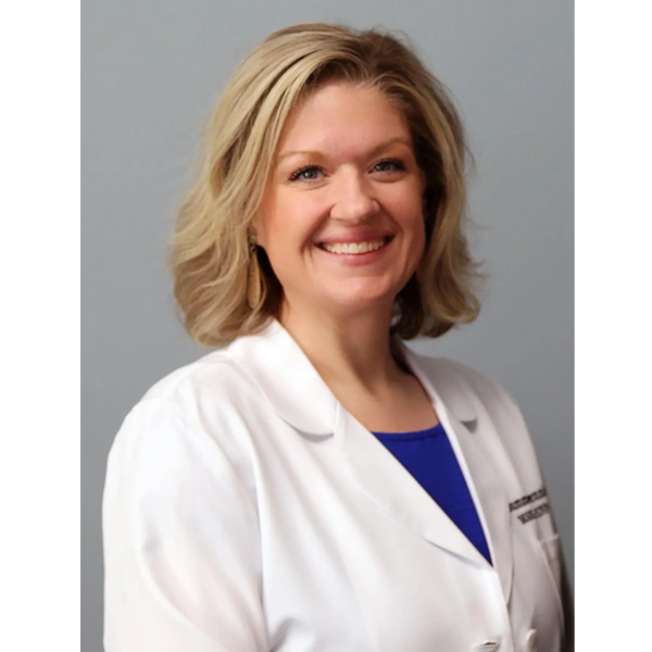 Smiling female doctor in a white coat with blonde hair against a gray background.
