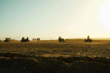 People ride ATVs across a dusty desert landscape during sunset.