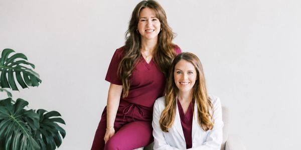 Two smiling female medical professionals in maroon scrubs and white coat.