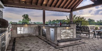 Modern outdoor kitchen with stone island and seating under a covered patio.