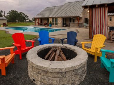 Colorful Adirondack chairs surround a stone fire pit near a pool at a countryside home.
