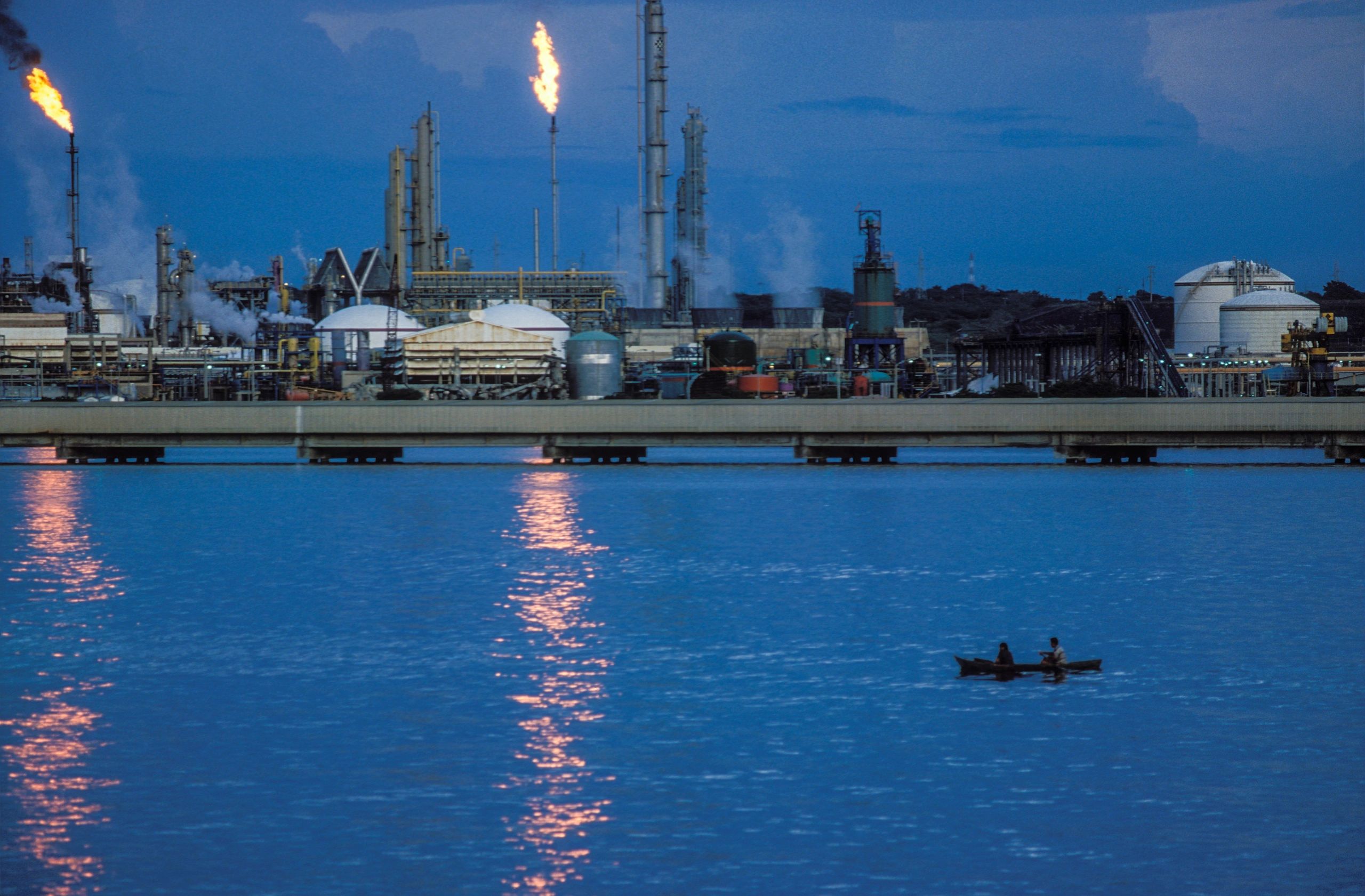 Industrial refinery with flames and a boat on water in foreground.