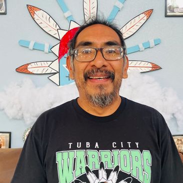 Smiling man wearing glasses and a Tuba City Warriors shirt.