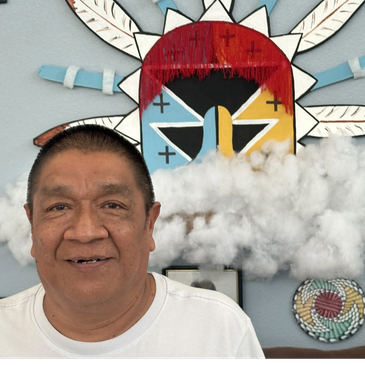 A man smiling in front of a colorful Native American mask and decorative wall art.