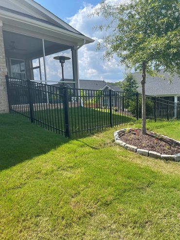 A backyard with a black metal fence, green grass, and a tree surrounded by stone edging.