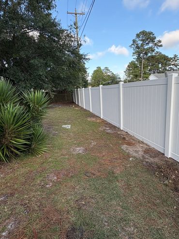 A backyard with grass, bushes, and a long white fence under a blue sky.
