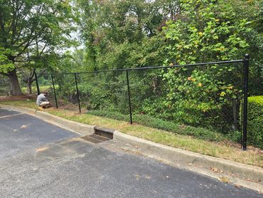 Man installing a black chain-link fence along a wooded area.