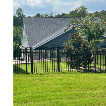 Black metal fence enclosing a green yard with trees and a house in the background.