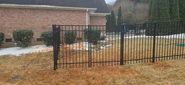 Black metal fence gate in a backyard with brown grass and a brick house.