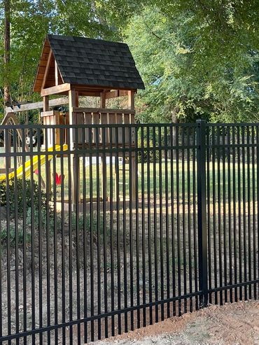 Black metal fence enclosing a backyard with a wooden playset and large tree.
