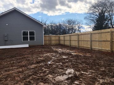 A muddy backyard next to a gray house with a wooden fence under a cloudy sky.