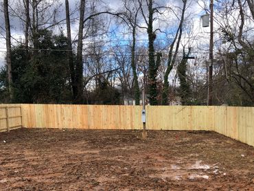 A muddy backyard enclosed by a new wooden fence with tall trees in the background.