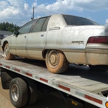 Old, dirty Chevrolet Lumina on a flatbed truck under a bright blue sky.