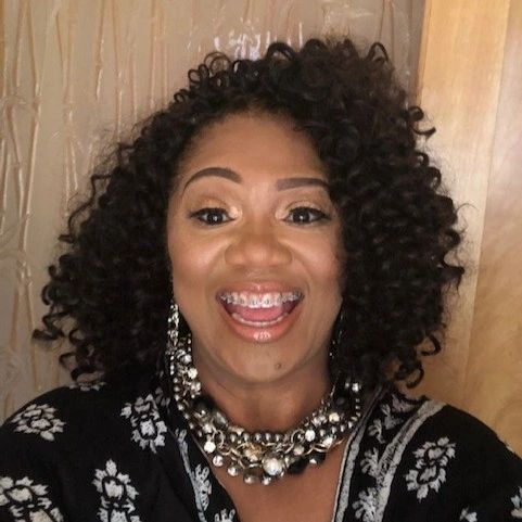 Smiling woman with curly hair and braces, wearing statement jewelry and floral attire.