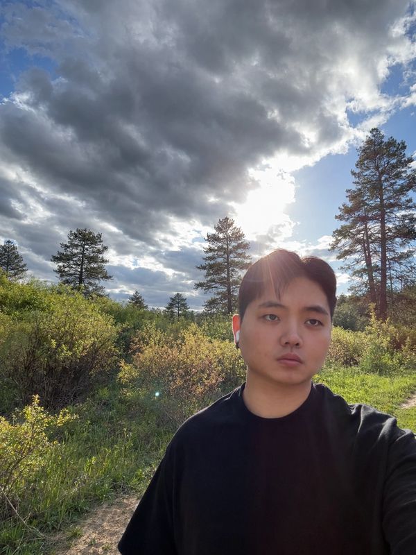 Young man taking a selfie outdoors with cloudy sky and sun.