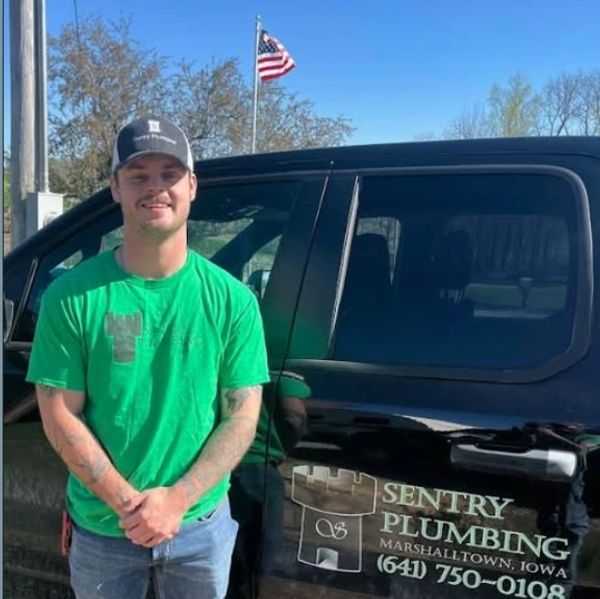A man in a green shirt stands next to a black Sentry Plumbing truck with an American flag in the background.